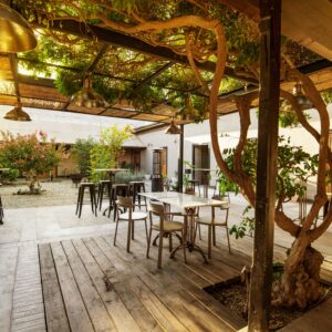 Patio interior con mesas y sillas, piso de madera, vegetación y pérgola de ramas, ambiente acogedor y natural al atardecer