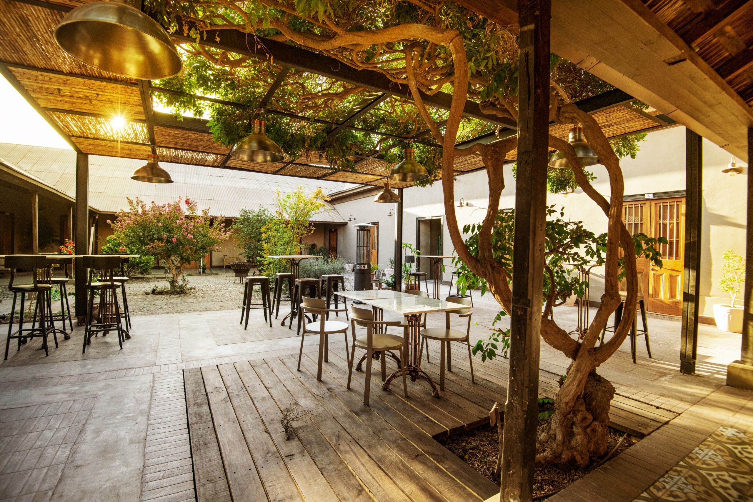 Patio interior con mesas y sillas, piso de madera, vegetación y pérgola de ramas, ambiente acogedor y natural al atardecer