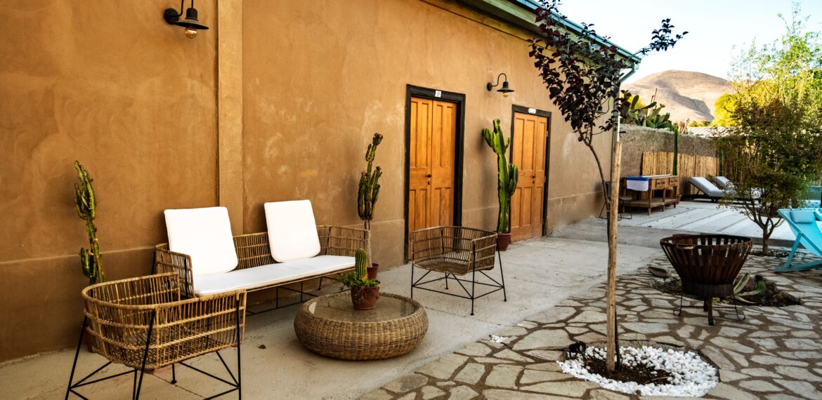 Zona exterior con sillones y mesa de mimbre, puertas de madera, cactus en macetas y ambiente tranquilo junto a un muro de color tierra. Se aprecian reposeras y vegetación en el fondo
