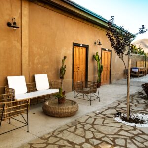 Zona exterior con sillones y mesa de mimbre, puertas de madera, cactus en macetas y ambiente tranquilo junto a un muro de color tierra. Se aprecian reposeras y vegetación en el fondo