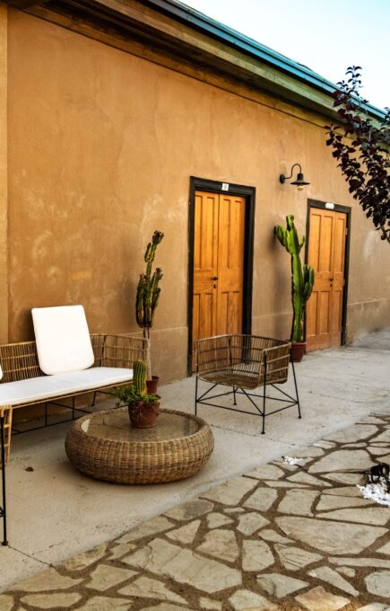 Zona exterior con sillones y mesa de mimbre, puertas de madera, cactus en macetas y ambiente tranquilo junto a un muro de color tierra. Se aprecian reposeras y vegetación en el fondo