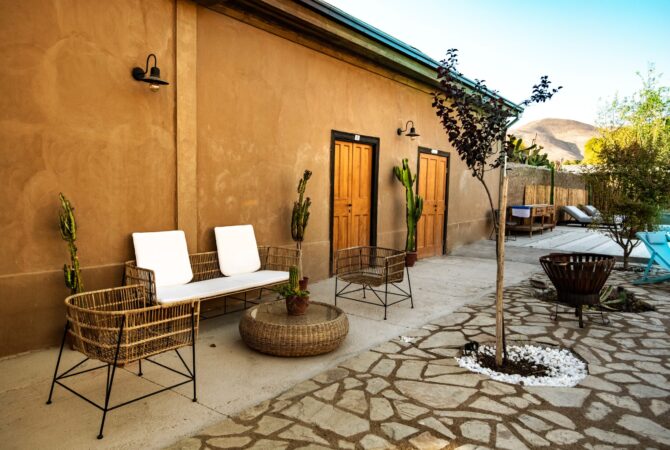Zona exterior con sillones y mesa de mimbre, puertas de madera, cactus en macetas y ambiente tranquilo junto a un muro de color tierra. Se aprecian reposeras y vegetación en el fondo