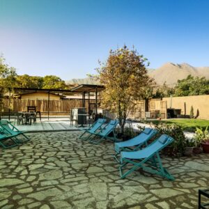 Patio exterior con reposeras azules, árboles y piso de piedra, rodeado de naturaleza y montañas al fondo