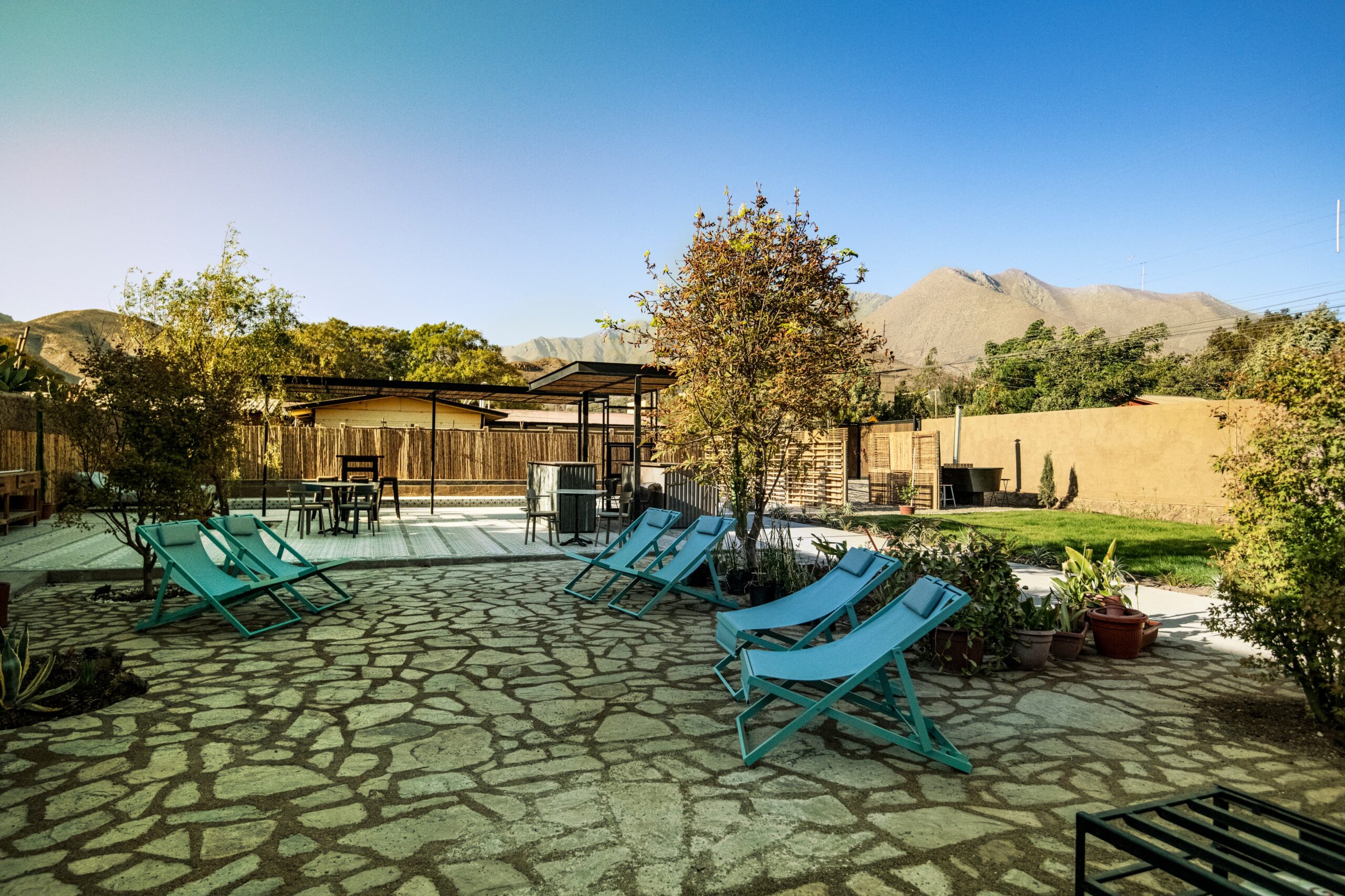 Patio exterior con reposeras azules, árboles y piso de piedra, rodeado de naturaleza y montañas al fondo