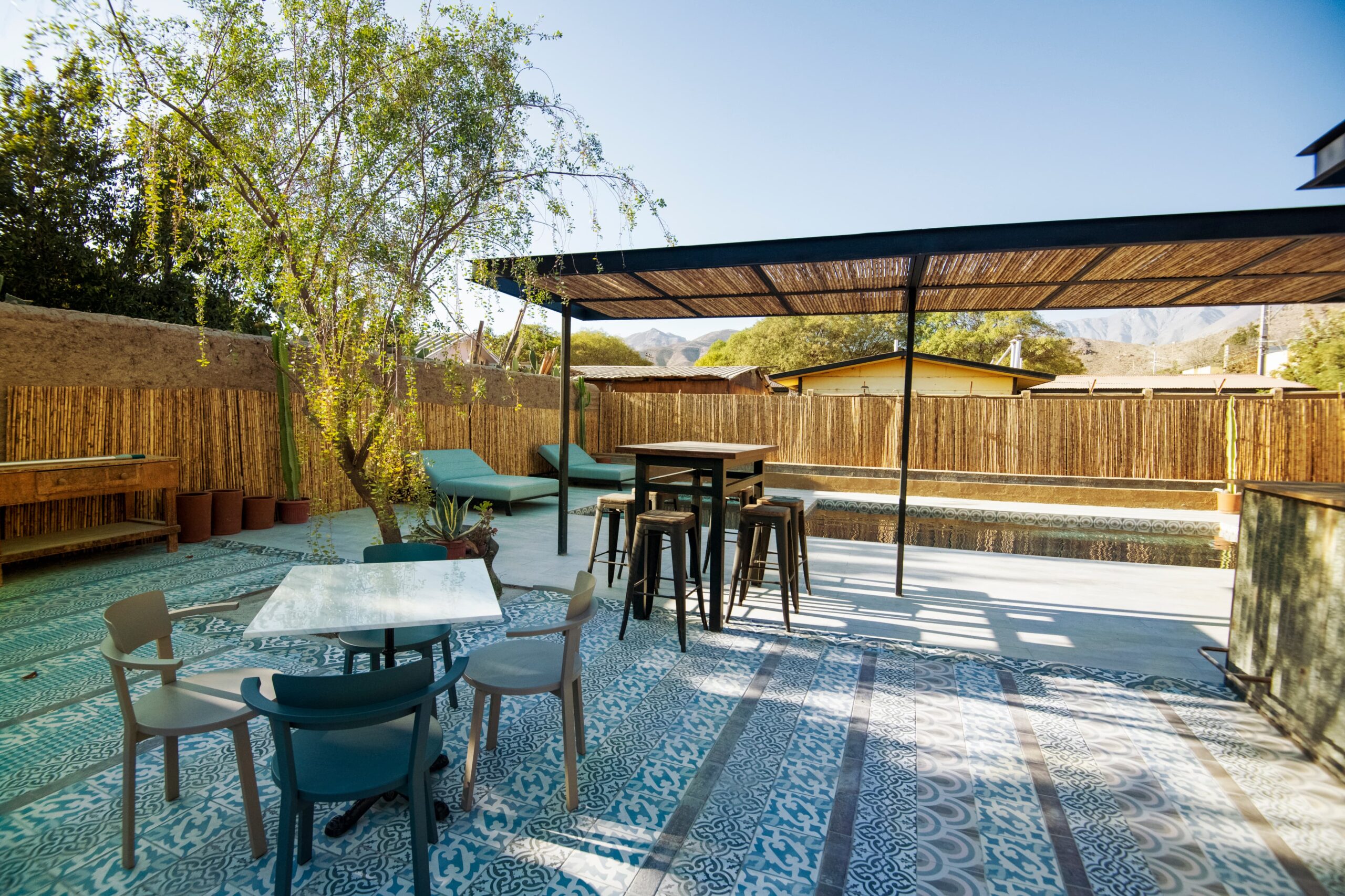 Terraza exterior con piscina, sillas y mesas bajo pérgola, rodeada de árboles y cercos de bambú, ambiente tranquilo y natural