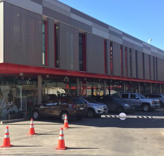 Vista exterior del centro comercial Paseo El Tamarugo mostrando el gimnasio Energy Fitness y la farmacia Cruz Verde, con autos estacionados y barrera de acceso al estacionamiento