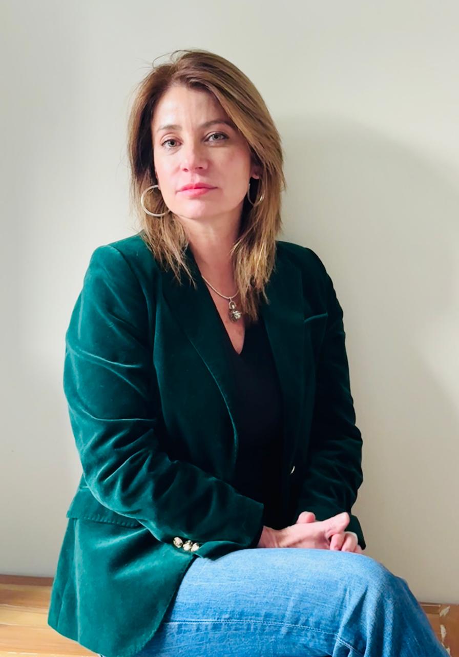 Retrato profesional de una mujer con chaqueta verde, sentada sobre una superficie de madera y fondo blanco, mirando a la cámara.