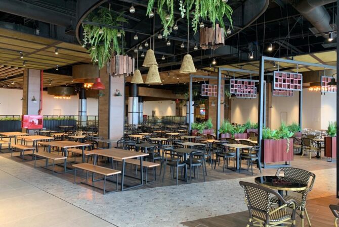 Área de comedor amplia y flexible, equipada con mesas grupales y bancas, ideal para food court o espacios de reunión. El diseño resalta por su iluminación cálida, abundante vegetación y detalles en madera, generando un ambiente acogedor y moderno.