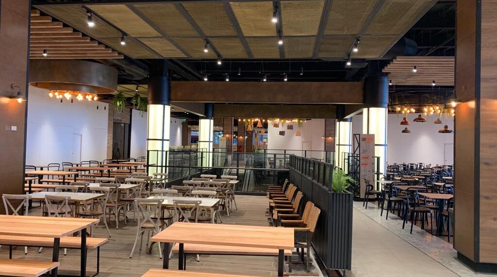 Comedor principal con mesas y sillas de madera y metal, iluminación cálida, plantas decorativas y detalles modernos en el diseño interior.