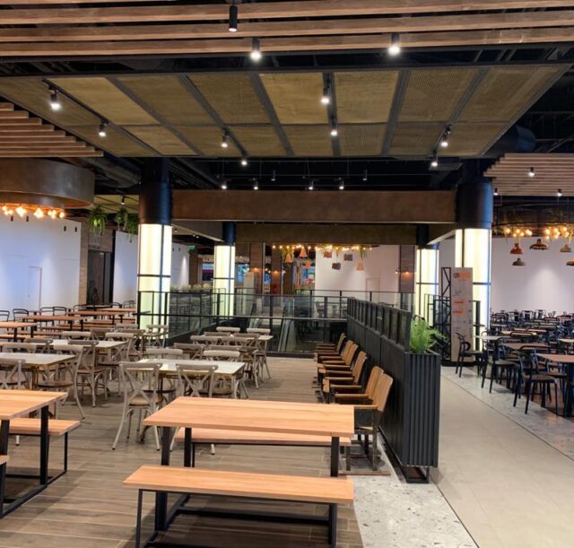 Comedor principal con mesas y sillas de madera y metal, iluminación cálida, plantas decorativas y detalles modernos en el diseño interior.