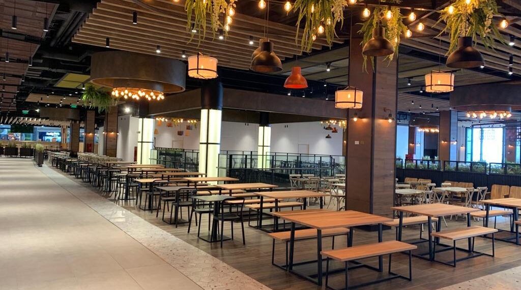 Vista de un amplio patio de comidas con mesas y bancas de madera, lámparas colgantes y plantas decorativas suspendidas, en un ambiente contemporáneo e iluminado.