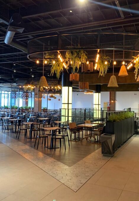 Espacio principal de un patio de comidas moderno, con mesas y sillas distribuidas alrededor de una estructura circular decorada con lámparas y plantas colgantes, junto a una escalera mecánica y columnas iluminadas.