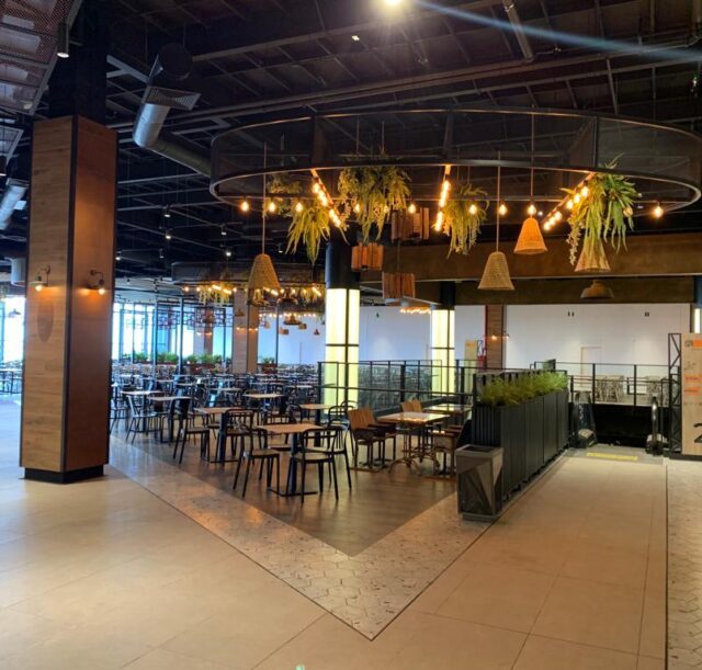 Espacio principal de un patio de comidas moderno, con mesas y sillas distribuidas alrededor de una estructura circular decorada con lámparas y plantas colgantes, junto a una escalera mecánica y columnas iluminadas.