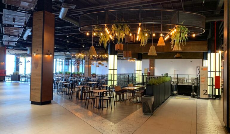 Espacio principal de un patio de comidas moderno, con mesas y sillas distribuidas alrededor de una estructura circular decorada con lámparas y plantas colgantes, junto a una escalera mecánica y columnas iluminadas.