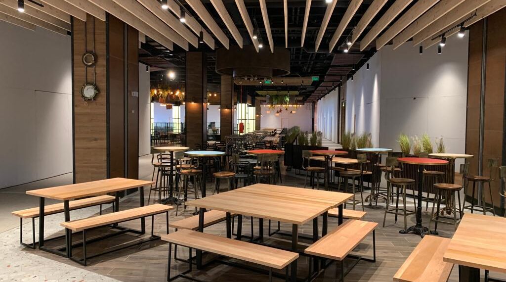 Área interior de un patio de comidas con mesas largas de madera, bancas y barras altas, bajo un cielo raso de vigas de madera y columnas revestidas, creando un ambiente cálido y moderno.