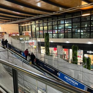 Imagen nocturna de un centro comercial con techo de madera y grandes ventanales. Se observa una rampa peatonal flanqueada por paneles protectores y señalización de la tienda Decathlon Viña del Mar. Varias personas caminan por el pasillo y se aprecian tiendas iluminadas y plantas colgantes decorativas.
