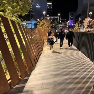 Personas caminando de noche por una pasarela iluminada frente al Mall Marina, con diseño moderno y jardineras a un costado.