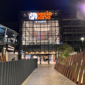 Vista nocturna del acceso al centro comercial Espacio Urbano 15 Norte, con iluminación moderna y personas transitando por la pasarela frontal.
