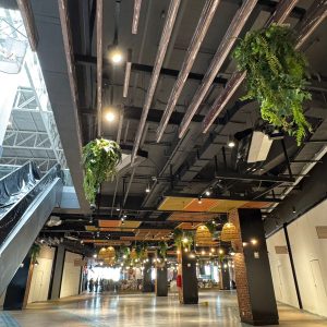 Vista amplia de un pasillo interior en un centro comercial. El techo tiene listones de madera y lámparas colgantes entre plantas decorativas. Se observa una escalera mecánica a la izquierda y pilares revestidos de ladrillo. Al fondo, hay luces cálidas y comercios, creando un ambiente acogedor y contemporáneo.