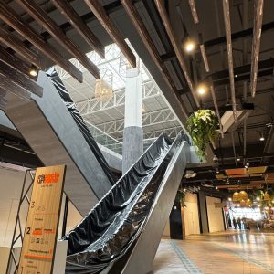 Primer plano de una escalera mecánica cubierta con plástico negro en el interior de un centro comercial moderno. En primer plano, un letrero de madera muestra información y servicios de “Espacio Urbano Las Rejas”. El techo presenta listones de madera y luminarias colgantes, junto con vegetación decorativa. Al fondo, el pasillo continúa con locales y luces cálidas.