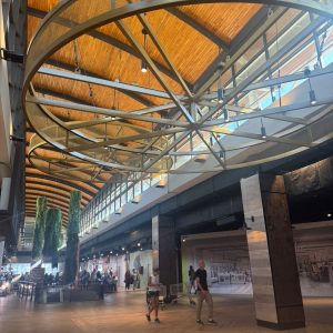 Espacio amplio y moderno dentro de un centro comercial, con techo de madera, estructura circular metálica en el techo, pilares revestidos y varias personas caminando y paseando con carros de supermercado. Se observan árboles interiores y vitrinas a los costados.
