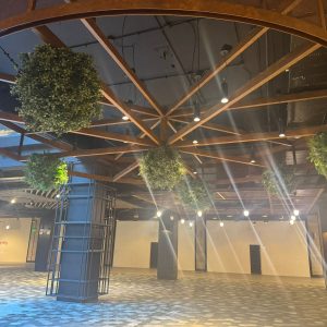 Detalle de un área interior del centro comercial con estructura circular metálica en el techo, esferas verdes colgantes y luces cálidas que crean un ambiente acogedor y moderno.