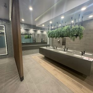 Lavamanos del centro comercial con grifería negra automática, espejo iluminado y decoración de plantas colgantes sobre estructura metálica, todo en un ambiente de tonos grises y madera clara.