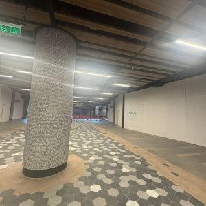 Vista de un pasillo amplio en un centro comercial en remodelación. El piso combina baldosas hexagonales en tonos grises y beige, con un patrón llamativo. Destaca una columna central revestida con pequeños mosaicos. El techo tiene listones de madera y luces lineales LED. Se observa una señal de salida verde y al fondo un acceso restringido con vallas rojas.