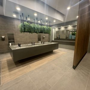 Vista amplia de lavamanos flotante con cubierta gris, grifería negra automática y vegetación suspendida, acompañada de paredes revestidas en porcelanato gris y detalles en madera decorativa.