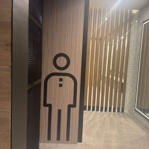 Acceso a baño de hombres en un centro comercial. Se observa un pictograma negro de figura masculina sobre panel de madera clara. El interior cuenta con separadores verticales de madera, paredes revestidas con azulejos grises y un espejo grande con iluminación LED. El piso es de cerámica beige. La ambientación es moderna y cálida.