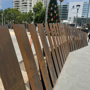Vista diurna de pasarela urbana delimitada por placas metálicas oxidadas inclinadas, con pavimento de piedra gris y un árbol de Navidad decorado al fondo, rodeado de edificios modernos y vegetación.