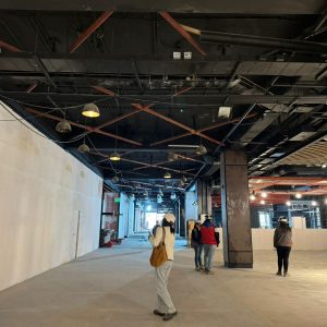 Personas con casco y equipo de seguridad recorren una zona interior en construcción de un centro comercial. El espacio muestra pilares, cielo raso negro con instalaciones y lámparas colgantes, además de muros de cierre provisorios. La iluminación es artificial y el ambiente luce amplio, con pisos y cielos aún en obra.