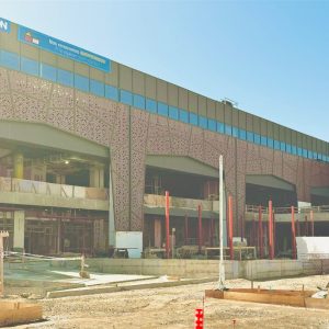 Fachada de un centro comercial en proceso de construcción, con andamios, paneles decorativos geométricos y ventanales superiores. Destaca un letrero azul de la tienda Decathlon en la parte alta del edificio. En primer plano, zona de obras y maquinaria de construcción bajo un cielo despejado