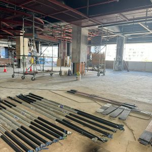 Vista de un área en construcción dentro de un centro comercial, con andamios metálicos, tubos y materiales de obra distribuidos sobre el suelo. Se observa el techo expuesto con tuberías, pilares y ventanas al fondo que dejan ver edificios residenciales en el exterior.
