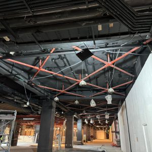 Vista del techo y parte del pasillo en construcción dentro de un centro comercial. Se observan estructuras metálicas de soporte pintadas de rojo, cables eléctricos, lámparas colgantes protegidas con plástico, y conductos eléctricos expuestos. También se ven andamios, columnas y una pared blanca sin terminar.