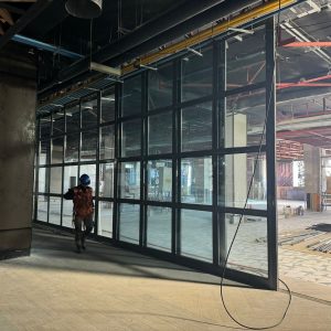 Obrero con casco y chaleco de seguridad caminando junto a una estructura de ventanales de vidrio y metal, en un sector interior en construcción de un centro comercial. Se observan elementos de la obra como andamios, materiales y cableado expuesto, además de tuberías y conductos eléctricos en el techo. El ambiente es industrial y de avance de obra gruesa.