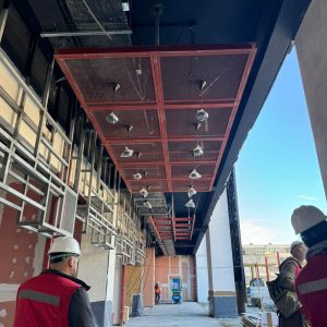 Personas con casco y chaleco de seguridad recorren un pasillo en construcción de un centro comercial. Se observa un cielo falso metálico rojo instalado, con luminarias cubiertas por bolsas de protección. A los lados se ven muros con terminaciones en proceso y estructuras metálicas para futuras instalaciones. Al fondo, más trabajadores y maquinaria elevadora. Es de día y parte del cielo está despejado.