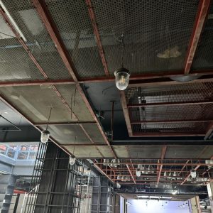Vista desde abajo de un cielo falso metálico de estructura roja con paneles tipo malla, instalado en el interior de un centro comercial en construcción o remodelación. Se observan luminarias colgantes con carcasa protectora y otras instalaciones eléctricas. La imagen muestra también parte de la estructura del edificio y sectores aún sin terminar.