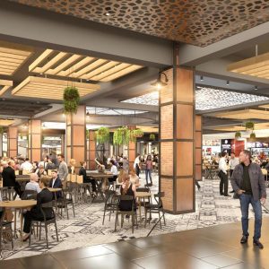 Área de patio de comidas en centro comercial con gran afluencia de personas, decoración en tonos cálidos, vegetación colgante, cielos falsos con listones de madera y lámparas industriales.