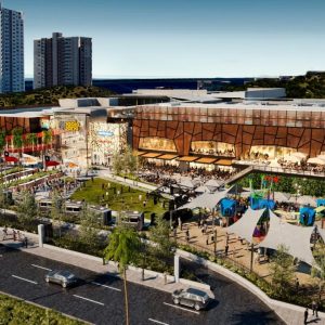 Render panorámico de un moderno centro comercial con edificios altos al fondo, espacios al aire libre con áreas verdes, food trucks, juegos infantiles cubiertos con toldos y terrazas de restaurantes en el segundo piso.