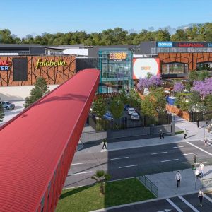 Vista exterior del centro comercial Espacio Urbano 15 Norte, con entradas señalizadas de Falabella y Sodimac, rodeado de áreas verdes, árboles con flores lilas, un pasillo techado rojo y peatones circulando por los cruces peatonales y veredas.