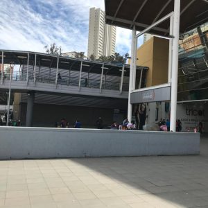 Vista del ingreso principal al centro comercial Espacio Urbano, con pasarela peatonal elevada, personas circulando y acceso a tiendas como Tricot