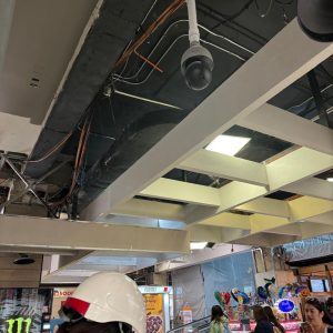 Vista de un área de centro comercial en proceso de remodelación, con el techo abierto mostrando cableado, cámara de seguridad y personas transitando junto a escaleras mecánicas y locales de comida