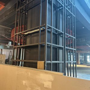Detalle de una estructura de metal negro alrededor de una columna en un centro comercial en remodelación, con paneles provisionales al frente y luz natural entrando por ventanas superiores.