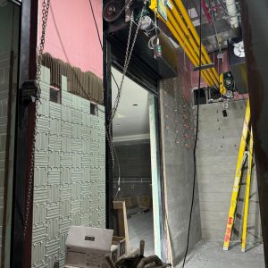 Vista de una zona en construcción con portón enrollable automatizado, cadenas, motores eléctricos y tuberías amarillas en el techo, junto a herramientas, cajas y una escalera amarilla dentro del recinto.