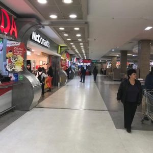 Pasillo interior de centro comercial con varios locales de comida rápida como McDonald's y Burger King, con personas caminando y clientes comprando comida.