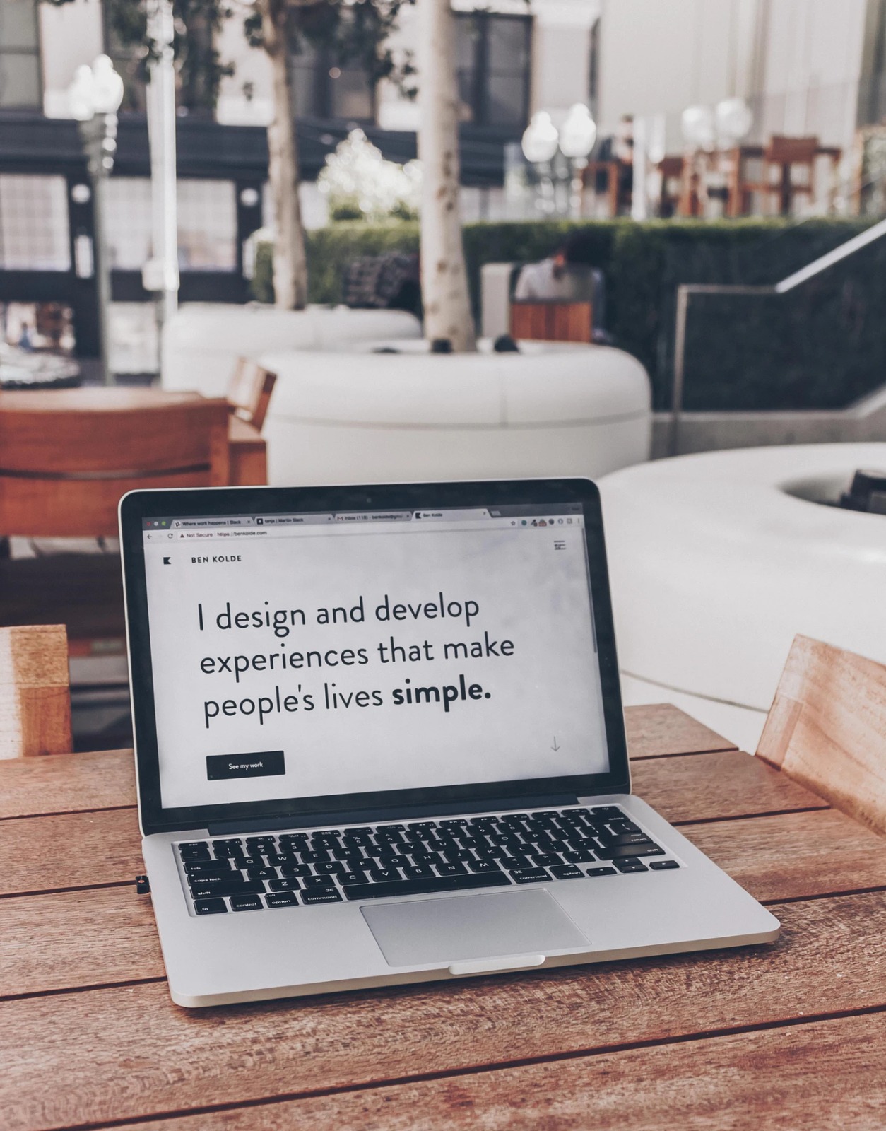 Notebook sobre mesa de madera mostrando el mensaje "I design and develop experiences that make people's lives simple" en un entorno de cafetería o cowork moderno con asientos y vegetación al fondo.