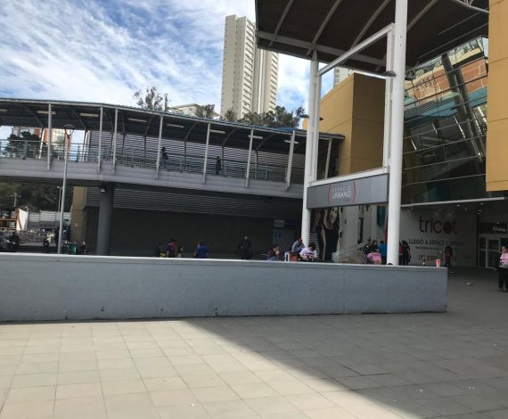 Vista del ingreso principal al centro comercial Espacio Urbano, con pasarela peatonal elevada, personas circulando y acceso a tiendas como Tricot